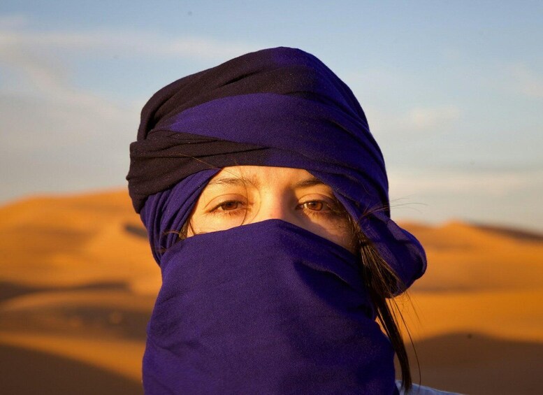A person in a purple garment and headwear, standing in a desert with sand dunes in the background. 2