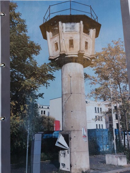 A weather station with a large cylindrical base, a protective canopy, and a building in the background. 2
