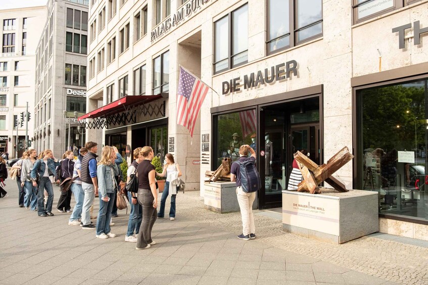 A group of people standing outside a building with a sign that reads 'DE MAUER.' 1