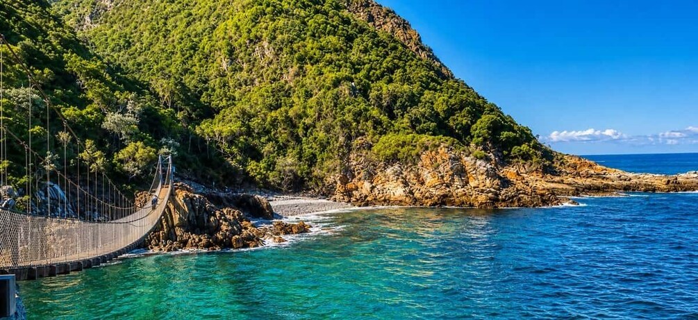 A suspension bridge over turquoise waters, a rocky coastline, and a forested hill. 5