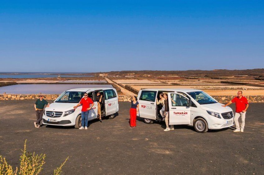 A group of people around a white van with 'TAMARAN' written on it, parked on a paved area with a clear sky and a barren landscape in the background. 4