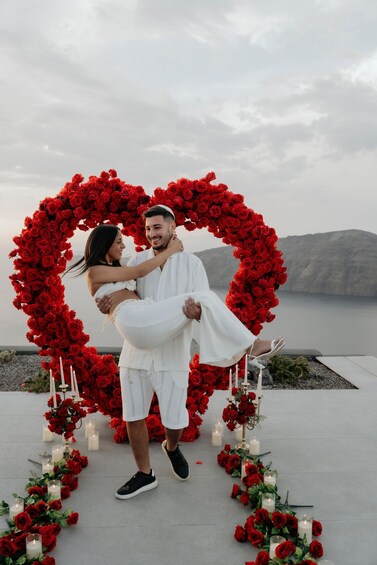 A couple embracing in a heart-shaped floral arrangement with candles, overlooking a body of water. 5