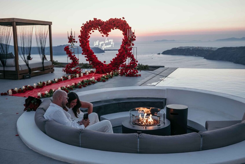 A couple sitting by a fire pit on a curved bench overlooking a body of water, with a heart-shaped floral arrangement and a canopy structure in the background. 3