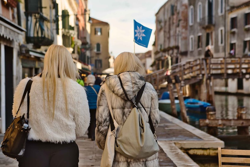 Two people walking down a canal-side street in a historic town, carrying bags. 2