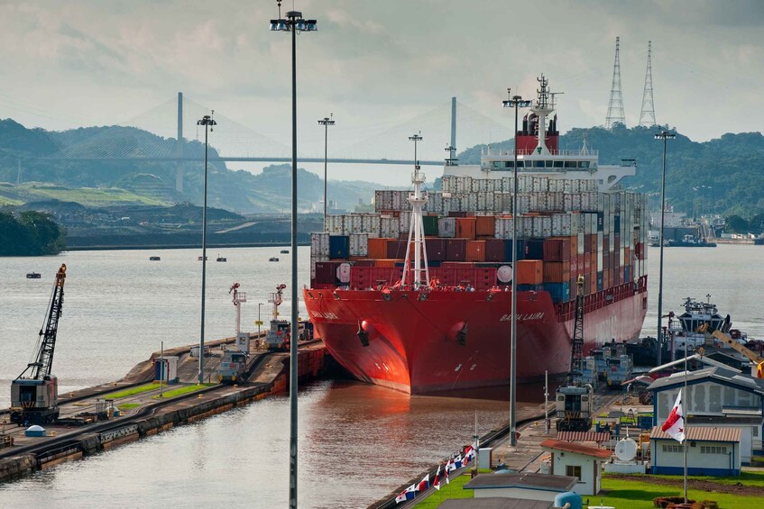 A large red cargo ship docked at a port with cranes and smaller boats in the background. 1