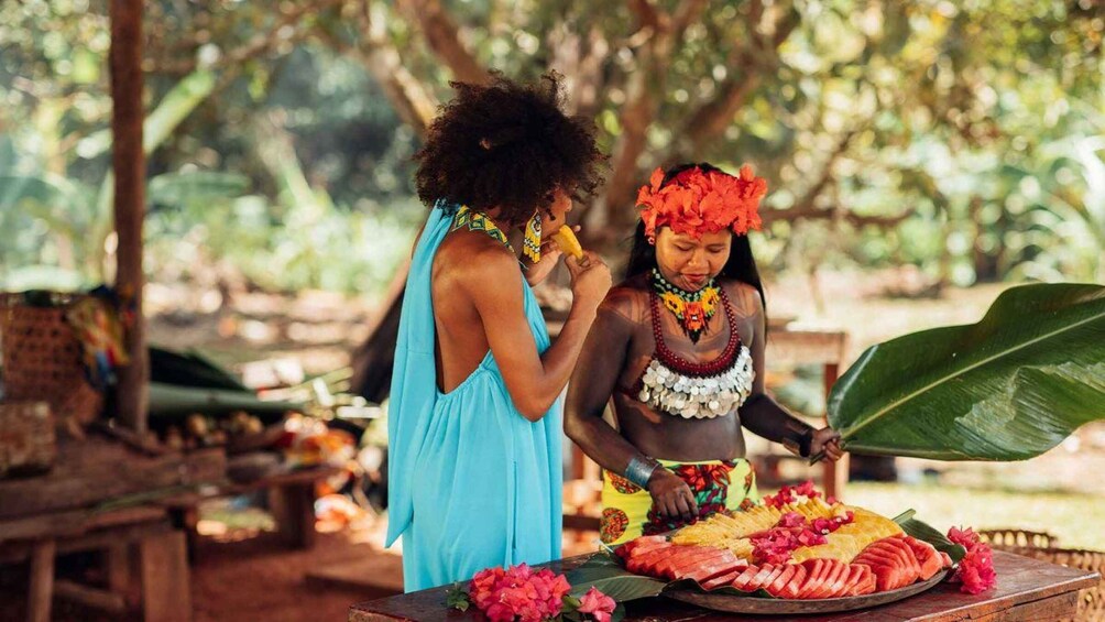 Two individuals in traditional attire, one holding a fruit, standing near a table with sliced fruits and flowers. 2