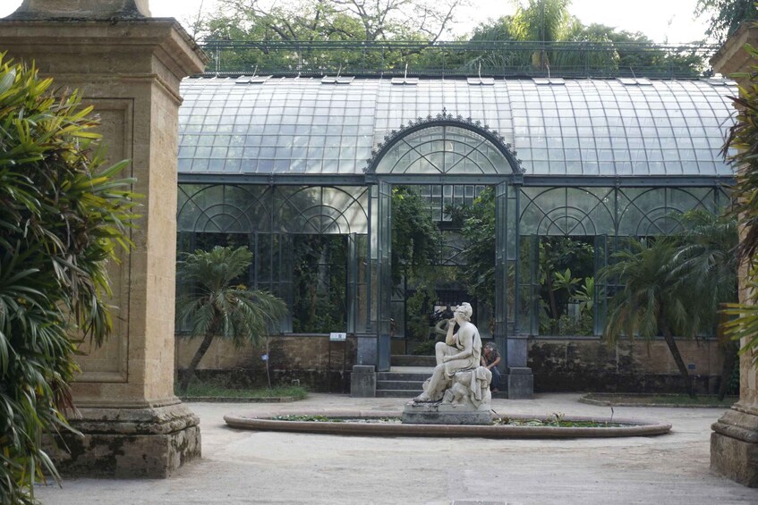 A classical statue in a garden with a greenhouse structure in the background. 1