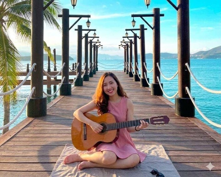 A person sitting on a wooden pier holding a guitar, with a clear view of the ocean and mountains in the background. 1