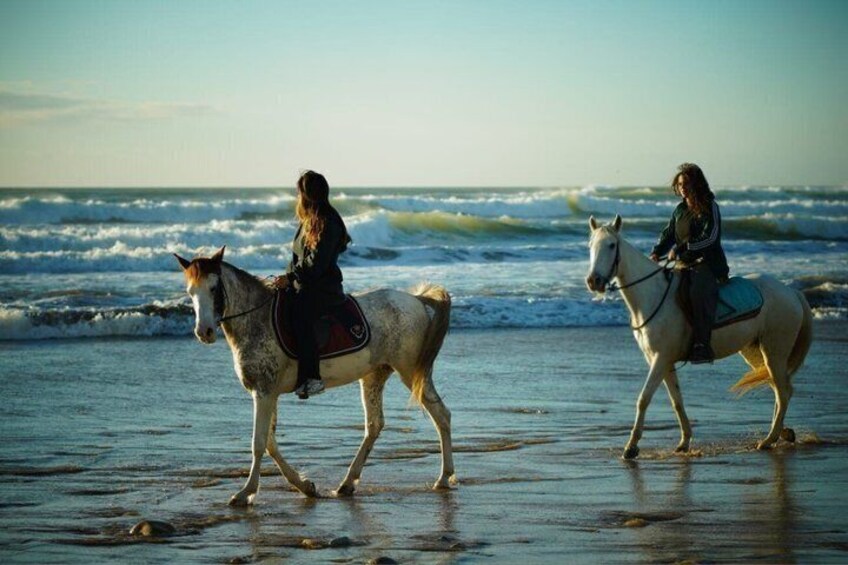 Two people riding horses on a beach with waves in the background. 1