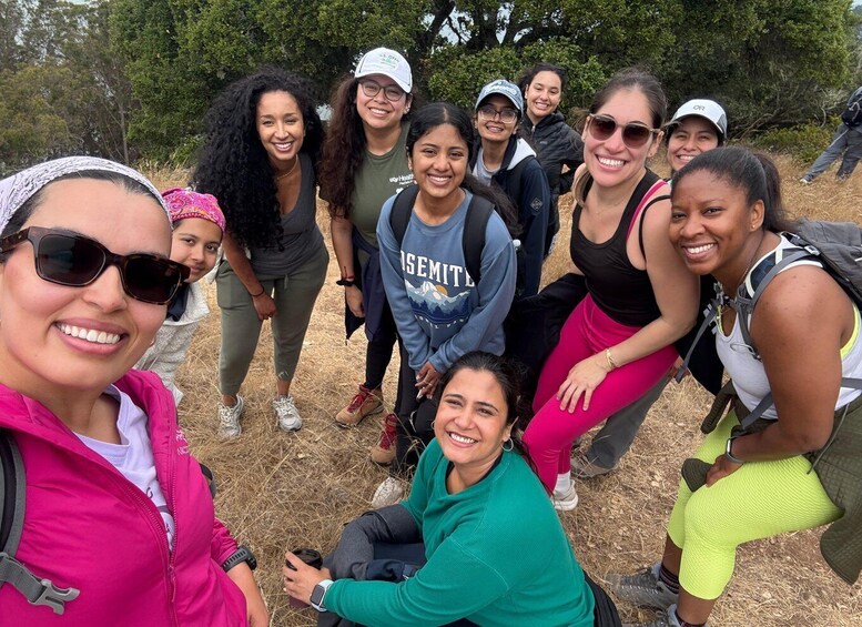 A group of people outdoors, some wearing casual clothing and backpacks, standing and sitting on dry grass with trees in the background. 3