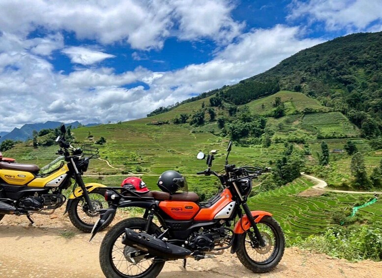Two motorcycles parked on a dirt path with terraced fields and a mountain in the background. 4
