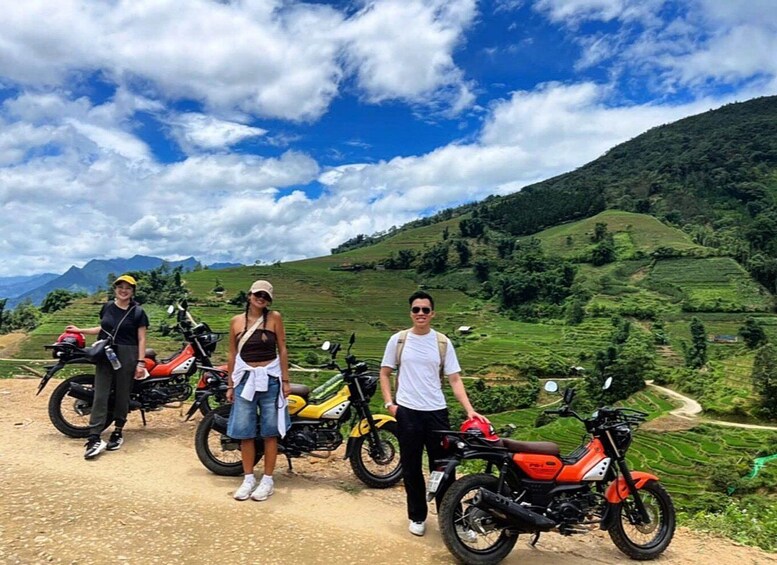 A group of people standing with motorcycles on a dirt path with green hills in the background. 3
