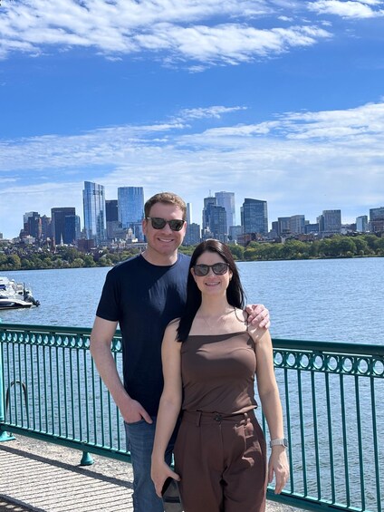 A couple standing on a waterfront promenade with a city skyline in the background. 1
