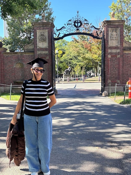 A person in graduation attire standing in front of a university entrance gate. 2