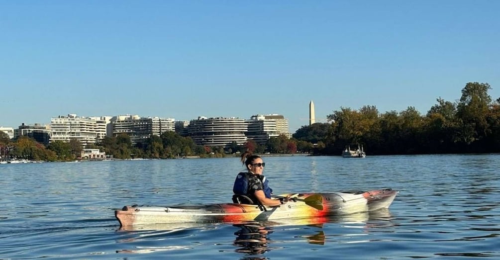 A person in a kayak on a calm body of water with a cityscape in the background. 1