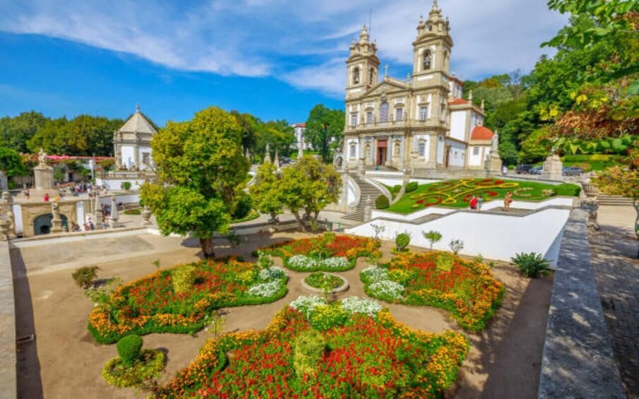 A historic building with two towers, surrounded by a well-manicured garden with various flower beds. 1