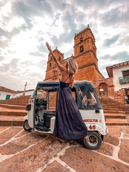 A woman in a long dress standing next to a white auto-rickshaw with the number 46, in front of a large brick tower. 1