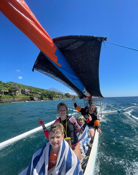 A sailboat with a blue and orange sail, people on board, and a coastal landscape with greenery and buildings in the background. 2