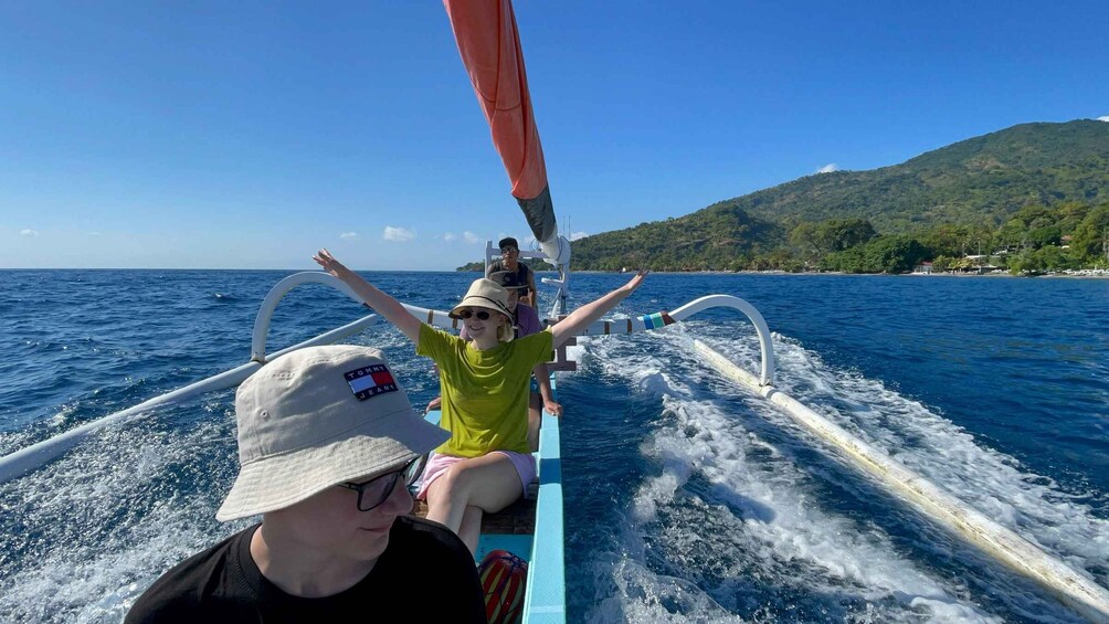 A boat with two people on board, one wearing a hat with a logo, and the other in a green shirt, with a mountainous coastline in the background. 3
