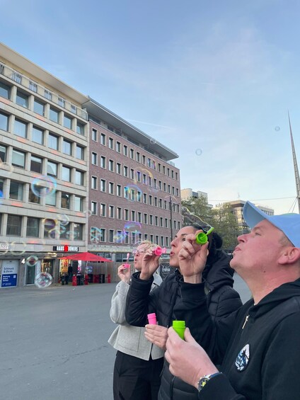 Three people blowing bubbles in an urban setting with buildings and a clear sky. 2