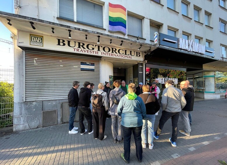 A group of people standing outside a building with a sign that reads 'BURGTORCLUB TRAVESTIE INTERNATIONAL.' 1