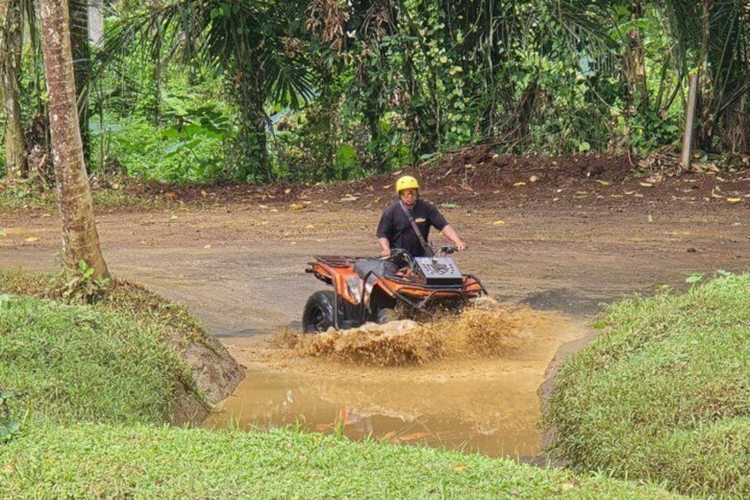 A person riding an ATV through a muddy water crossing in a tropical setting. 5