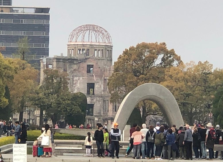 A crowd of people gathered around a large arch monument in a park with a historic building in the background. 1