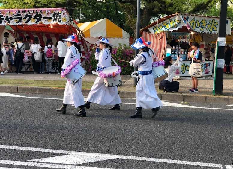A street parade with performers in white costumes and hats, marching down the road. 2