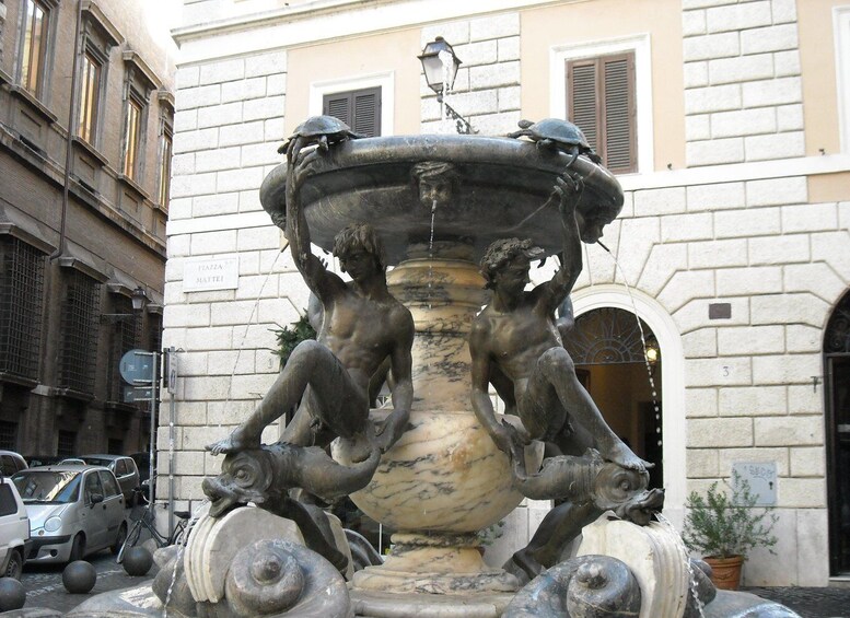 A classical fountain with two statues, a building with a sign reading 'FLAZA MATTEI,' and parked cars. 3