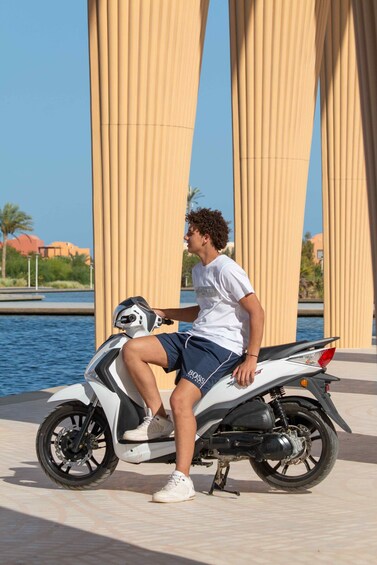 A person sitting on a white scooter with a helmet, near a body of water and a building with vertical columns. 5
