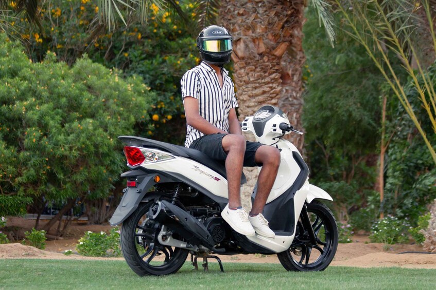 A person sitting on a white scooter with a helmet, in a garden with palm trees and yellow flowers. 3