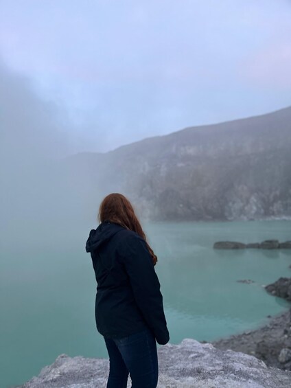A person standing on a rocky outcrop overlooking a turquoise body of water with mist in the background. 5