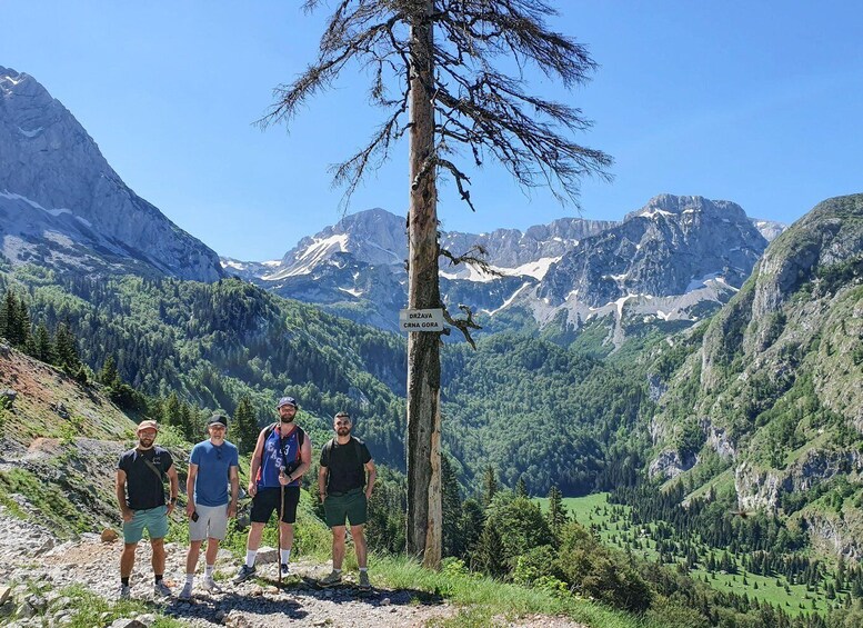 Four hikers on a mountain trail with a sign indicating 'DRZAVA CRNA GORA' and a helicopter flying in the background. 5