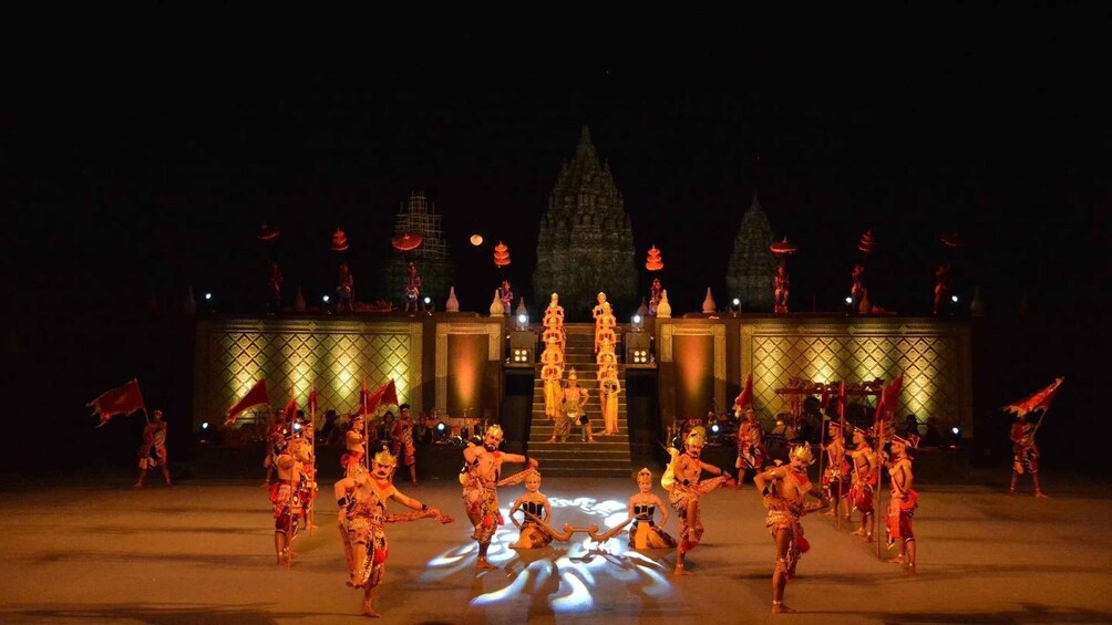 A nighttime cultural performance with dancers in traditional costumes, a temple-like structure in the background, and a large illuminated lotus flower on the ground. 2