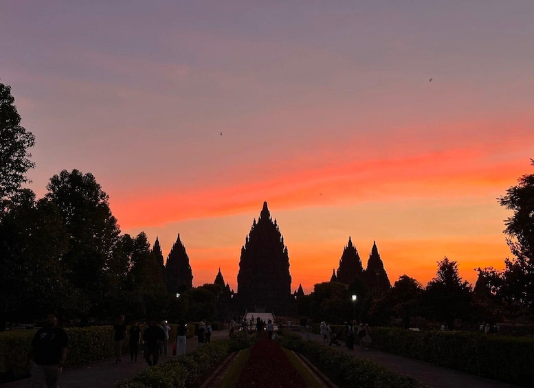 A temple complex with a row of tall spires, surrounded by trees and a pathway with people walking. 1