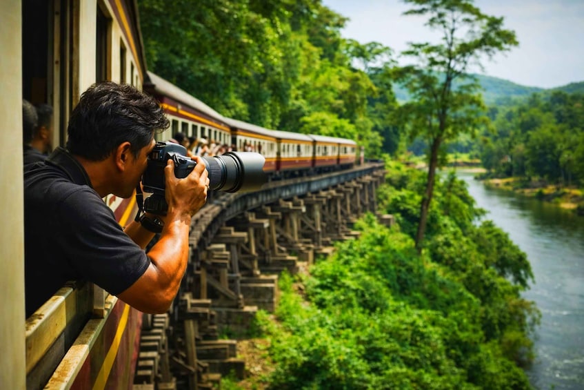A person taking a photograph of a train on a bridge with a river and greenery in the background. 1
