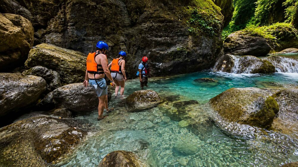 Three people in a clear stream surrounded by large rocks and greenery. 4