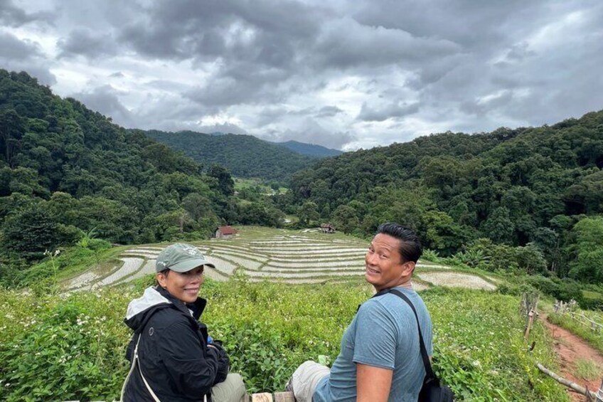 Two people sitting on a grassy hill overlooking terraced fields and a forested valley. 5