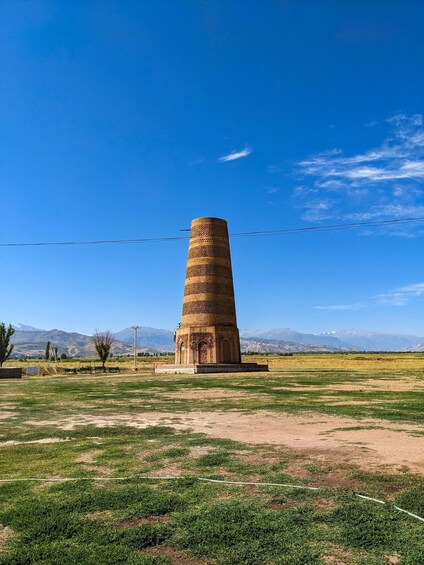 A tall, cylindrical tower with intricate brickwork, set against a backdrop of mountains and a clear blue sky. 3