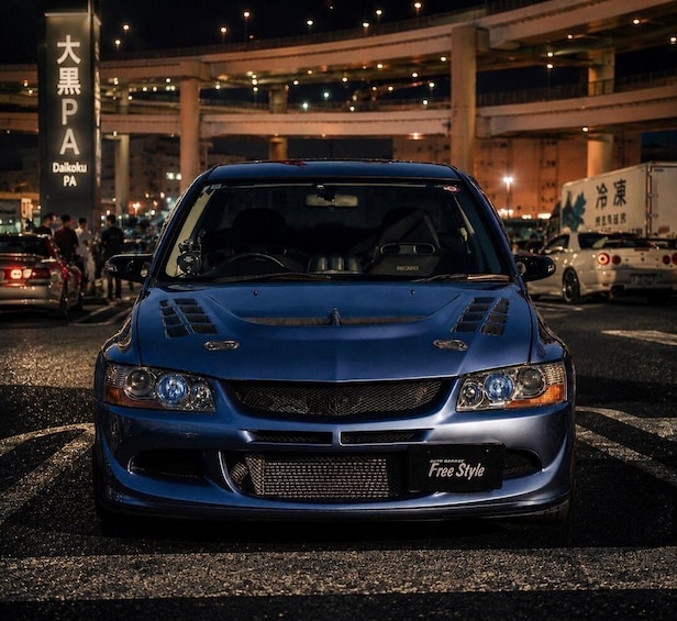 A blue car parked on a street at night with a sign reading 'Daikoku PA' in the background. 1
