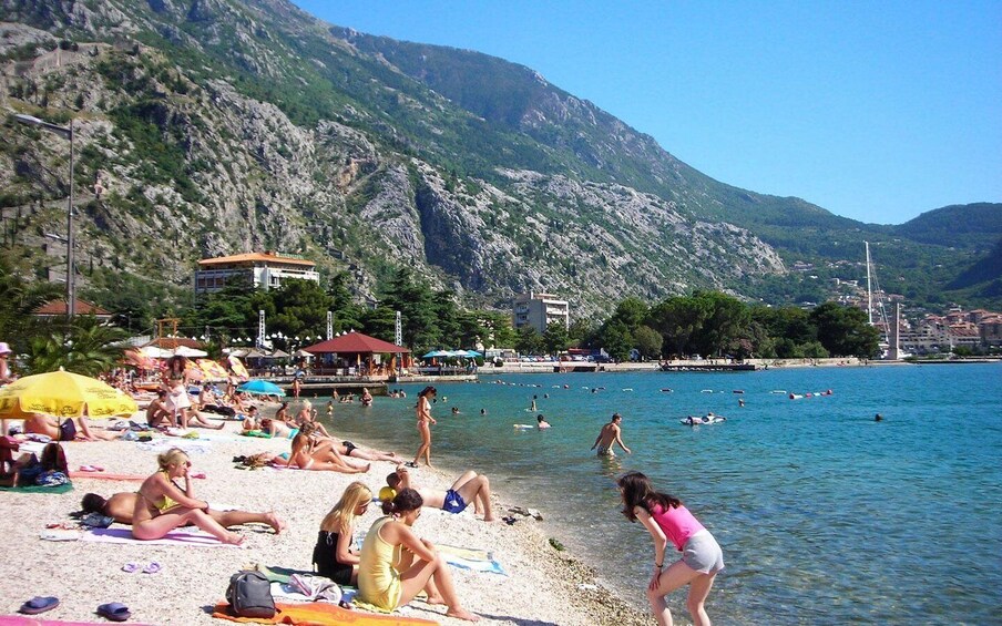 A beach with people sunbathing and swimming, surrounded by mountains and buildings. 2