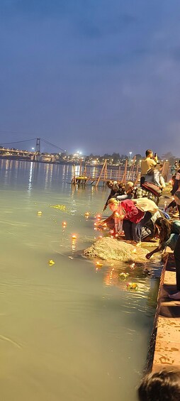 A river with people performing rituals, a bridge in the background, and a calm evening setting. 2