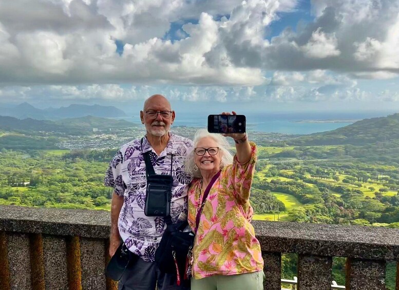 Two people standing on a stone platform with a scenic view of a coastal landscape and mountains in the background. 3