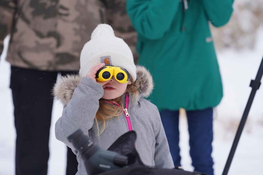 A person wearing a white beanie and yellow goggles, standing outdoors in a snowy environment. 5