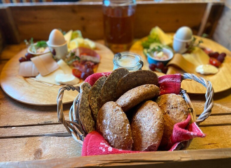 A wooden table with a basket of bread, a cheese board with various cheeses and accompaniments, and a glass of beverage. 4