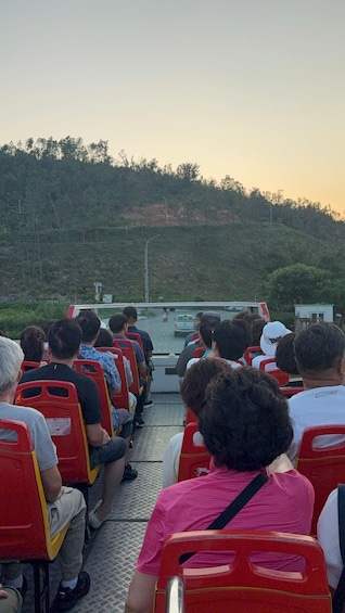 People seated on red chairs facing a bus on a road with a hilly landscape in the background. 2