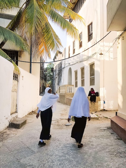 A narrow alleyway with two women walking, palm trees, and a building with barred windows. 2