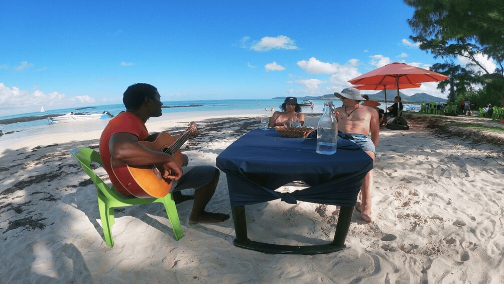 A man playing guitar at a beachside table with a drink, under a red umbrella. 4
