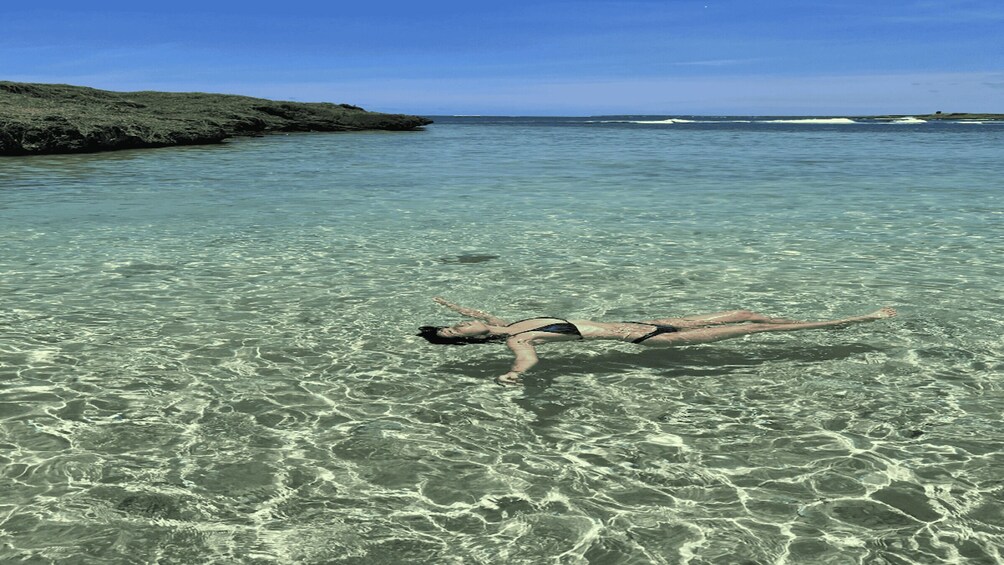 A person floating on a clear shallow sea with rocky coastline in the background. 5