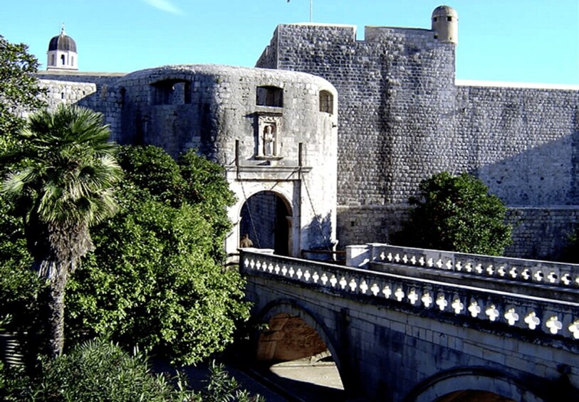 A stone bridge with a decorative railing, a stone wall with a tower, and palm trees. 2